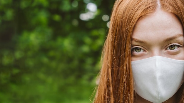 New normal outdoor portrait of redhead woman with protective mask on green trees backgroung young