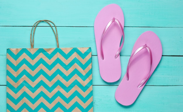 青い木製の背景に新しいピンクのビーチサンダルと紙袋。ショッピングのコンセプト。上面図