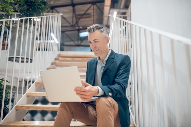 New project. Cheerful successful adult man sitting on stairs working on laptop confidently looking at screen in illuminated office space