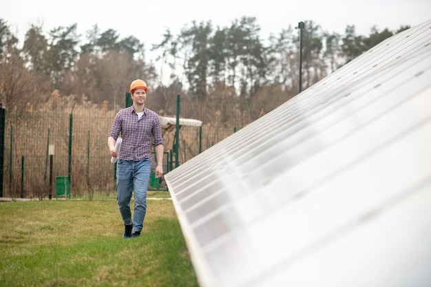 New project. Young adult man in safety helmet with drawing in his hand walking near solar panel in countryside