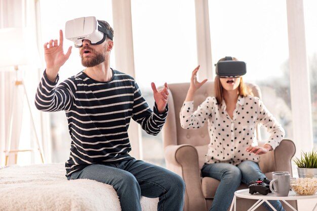 Новая реальность. Довольно эмоциональный приятный мужчина протягивает руку, пробуя гарнитуру VR и сидя на кровати