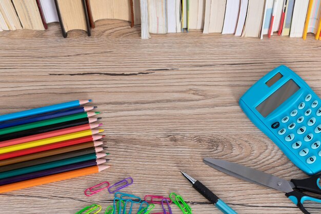 The new school and office supplies lie on a wooden desk top next to a row of books