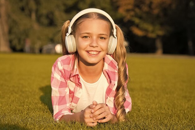 子供の幸せな子供時代の思い出のための新技術 学校に戻って音楽を聴く 子供は公園で勉強します ヘッドフォンで緑の芝生でリラックス 小さな女の子はオーディオブックを聞きます