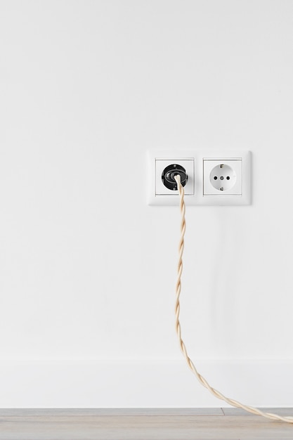 Photo new white plastic european electrical outlet isolated on white plastered wall. an electrical plug with a vintage cable is plugged into an outlet. gray wood floor. copy space