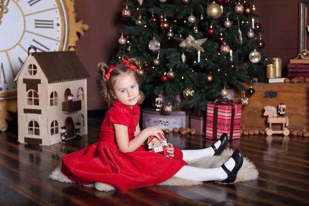 New Year 2020. Merry Christmas, happy holidays. Little girl in a red vintage dress sits near a decorated Christmas tree with a wooden toy The Nutcracker. Family holiday. Happy kid enjoys the holiday.