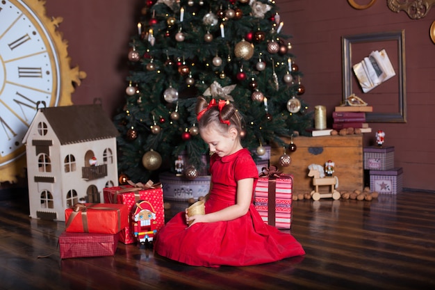 Новый год 2020. Счастливого Рождества, веселых праздников. Портрет маленькой девочки с свечой. Маленькая девочка держит свечу в ее руках перед рождественской елкой и подарками. Рождественский декор для дома, новогодняя комната
