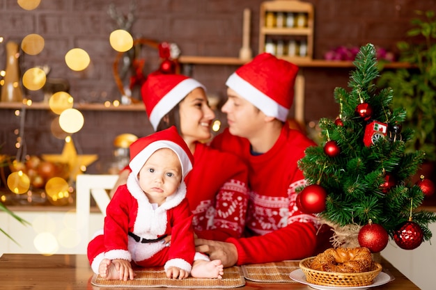 New Year or Christmas, close-up of a baby in a red sweater and Santa Claus hat in the kitchen, a happy young family mom, dad and baby at the Christmas tree smiling, hugging