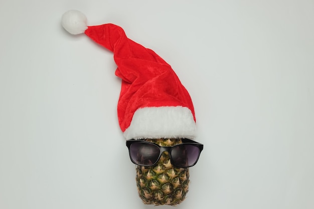 Photo new year, christmas concept. pineapple dressed in glasses with santa hat on white background. top view.