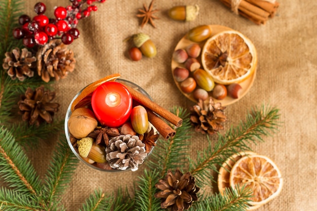 Capodanno e decorazioni natalizie di bacche, coni, noci, fette d'arancia in una composizione con una candela rossa sullo sfondo di tela da imballaggio.