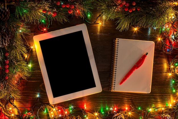 新年とクリスマスのモックカップ。木製の背景、テキスト用のスペース、コピースペースにクリスマスライトが付いたノートブック。上から。休日の冬の計画の概念。フラットレイスタイル。