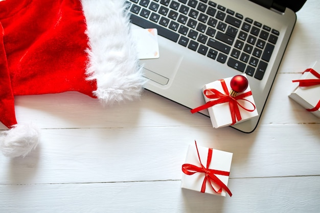 Acquisti online di capodanno e natale a casa, concetto di quaranina, notebook, carta di credito, cappello da babbo natale e confezione regalo su fondo di legno bianco, vista dall'alto, spazio di copia