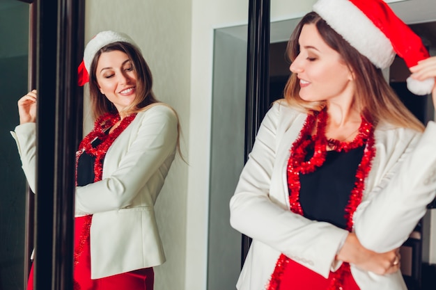 Новогодняя или рождественская вечеринка моды. Женщина проверяет одежду, смотрит в зеркало, носит шляпу Санты