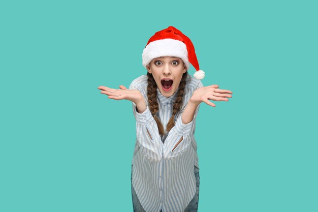 New year concept. amazed beautiful girl in striped light blue shirt in red christmas cap standing and looking at camera with raised arms and surprised face. studio shot isolated on green background.