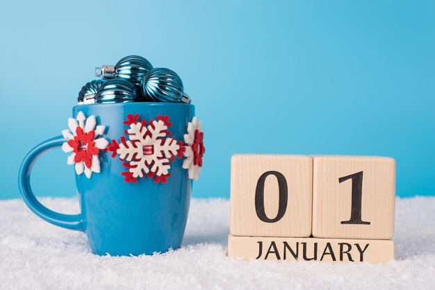 New Year concept. Close up photo of blue coffee cup full of small baubles and wooden cubes calendar with New Year date standing in fluffy white snow on blue background