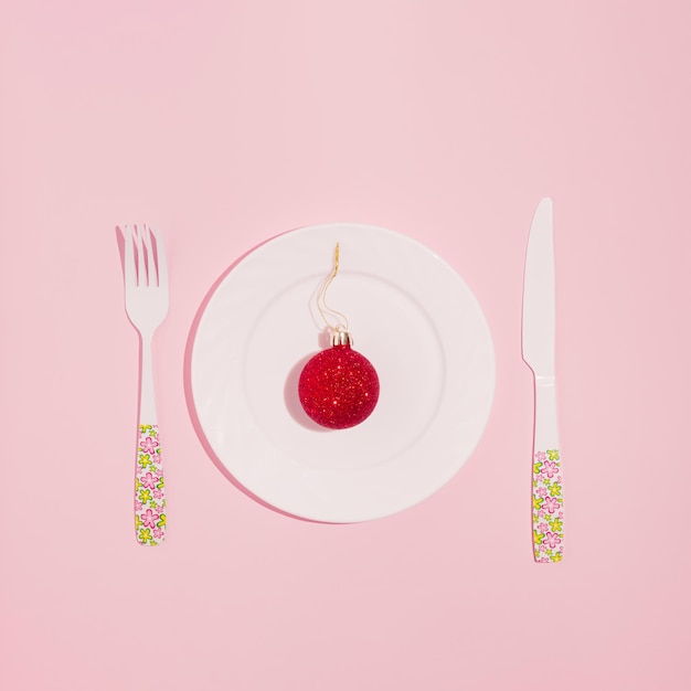 New Year creative layout with plate, fork, knife and red Christmas bauble decoration. Pastel pink background. Holiday party dinner idea.