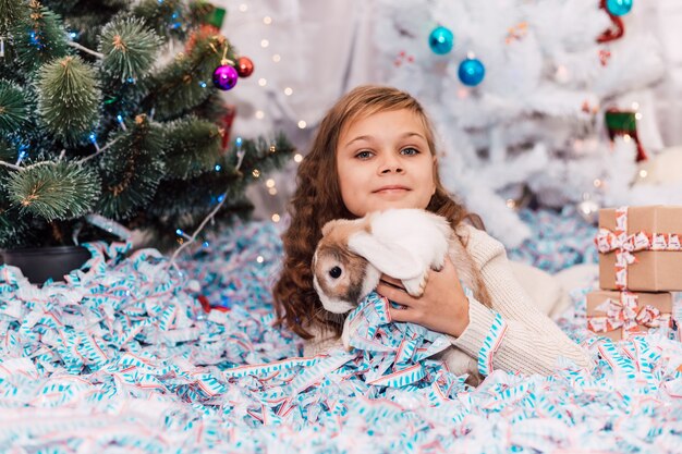 New year elegant girl lies on her stomach near a green decorated spruce holding a rabbit