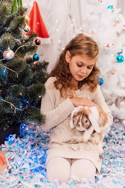 New year elegant girl in a light dress near a green decorated spruce holding a rabbit