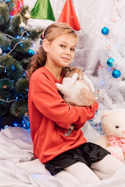 New Year a girl in a New Years sweater holds a rabbit near the Christmas tree