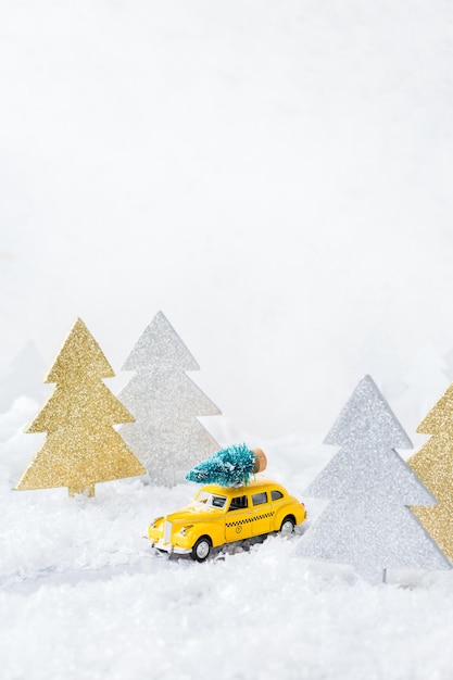 New year holiday concept Yellow toy taxi car carrying christmas tree in a snow forest Copy space greeting card with festive decoration