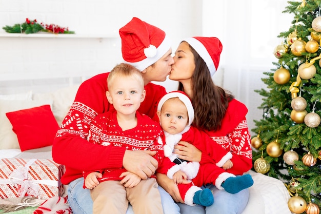 Фото Новый год или рождество, счастливая семья с двумя детьми на кровати дома у елки в красных свитерах улыбается, обнимается и целуется, поздравляя с праздником