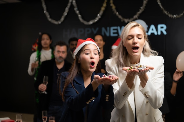 Photo new year party group of young people in santa hats blowing confetti while celebrating new year