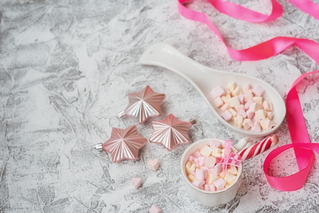 New year's or christmas composition cup with marshmelow with candy among new year's, shiny toys and pink ribbon on a bright textural background. flat lay, layout