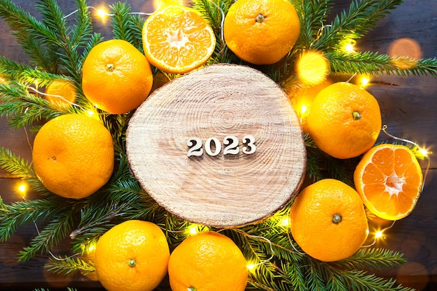Фото Новогодний праздничный фон на круглом срезе дерева, окруженном мандаринами, живыми еловыми ветками и золотыми огнями гирляндами, с деревянными числами, датируемыми 2023 годом. цитрусовый аромат, рождество. место для текста.