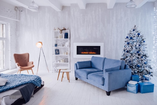 New Year's interieur van het appartement. Grote ramen, blauwe bank, kunstmatige open haard, bed, verlichting, witte en blauwe kerstboom, fauteuil en stijlvolle vloerlamp. Loft-stijl. Betonnen muur. Gezellig huis.