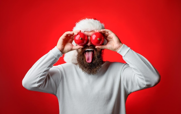 New Year toy. Happy Santa hold christmas balls. Bearded man holds Christmas toy near eyes. Christmas mood. Happy holidays. Merry Christmas. New Year. Santa's hat. Funny faces. Santa shows tongue.