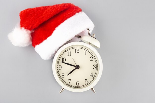 New Years or Christmas creative concept white alarm clock in a Christmas hat on a gray background