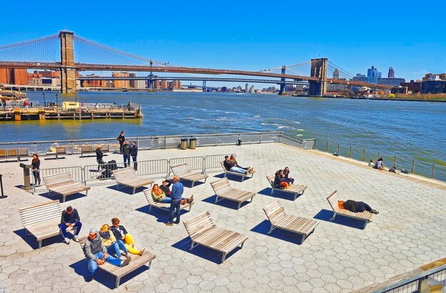 New York, USA - April 25, 2015: Tourists relaxing on sunbeds in Pier in Lower Manhattan, New York. View from Ferry on Pier, Brooklyn Bridge and Manhattan Bridge and Brooklyn, USA, East River