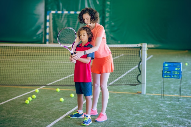 Newbie. Female coach in bright clothes teaching a boy to play tennis