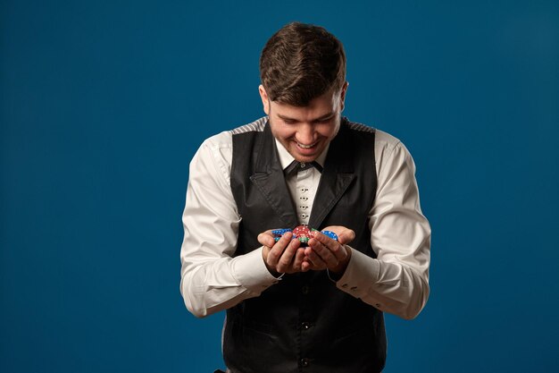 Photo newbie in poker in black vest and white shirt holding some colored chips posing against blue backgro