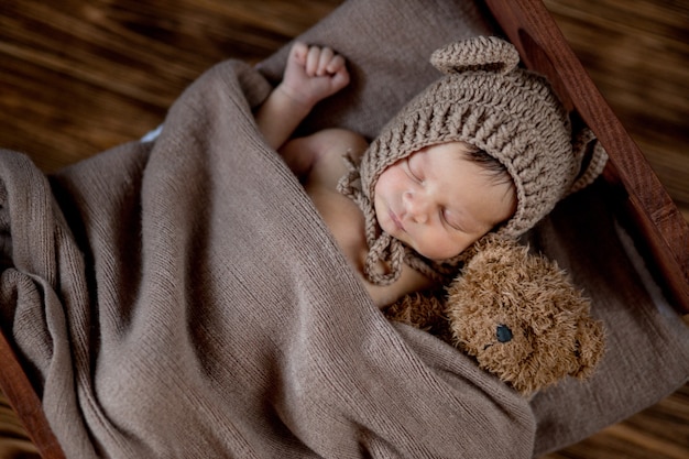 Newborn baby, beautiful infant lies and holding a tiny teddy bear in the bed on wooden surface.