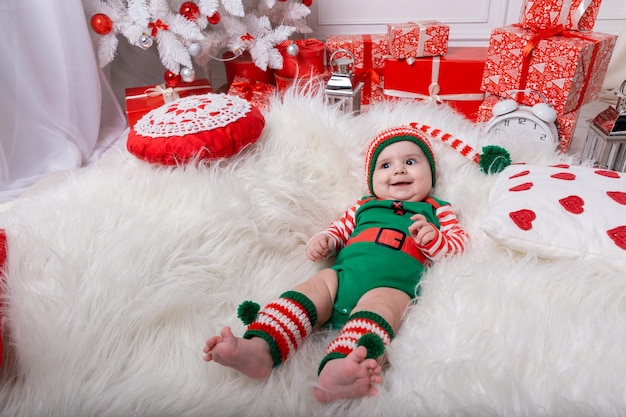 Newborn baby boy dressed in gnome costume lying on white fur carpet among christmas decorations