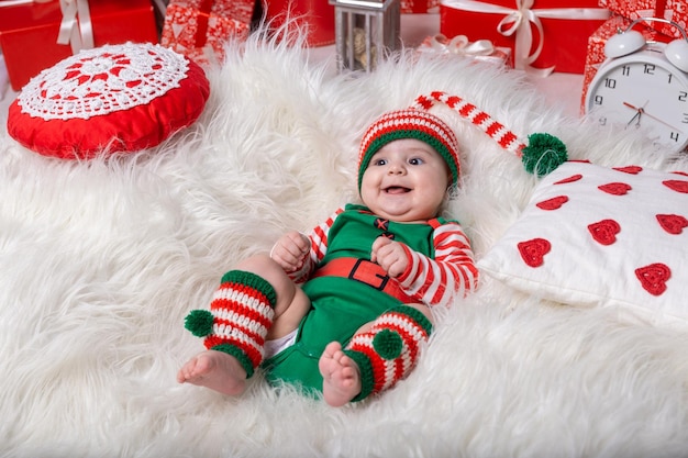 Newborn baby boy dressed in gnome costume lying on white fur carpet among christmas decorations