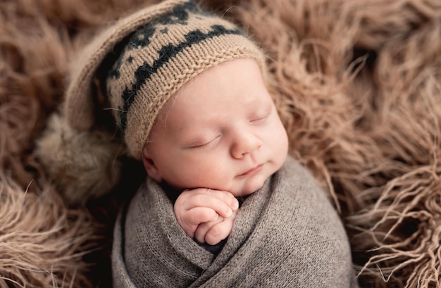 Newborn baby boy swaddled in fabric sleeping and holding his hands close to cheeks. Infant kid napping on fur