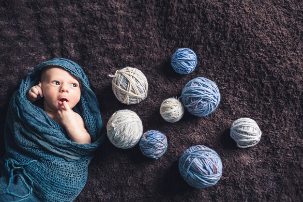Newborn baby lies wrapped in blanket amid tangles of thread and looks away.