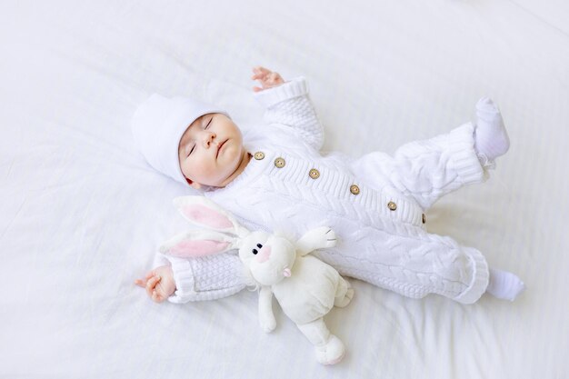 A newborn baby sleeps in a crib in a white warm jumpsuit and a hat on a white cotton bed with a soft toy healthy baby sleep