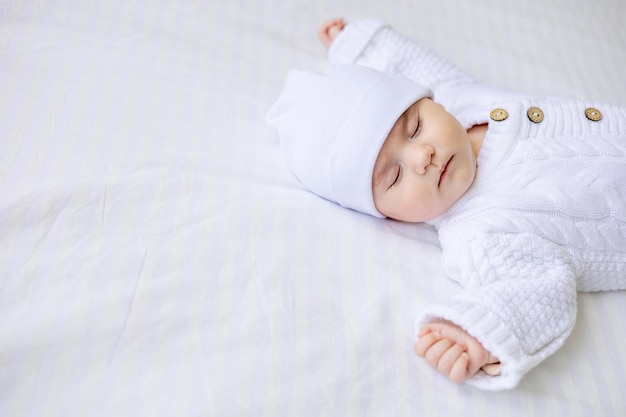 갓난 아기는 흰색 따뜻한 점프수트를 입은 유아용 침대에서 잠을 자고 흰색 절연 면 침대에 모자를 쓰고 건강한 아기 수면을 닫습니다