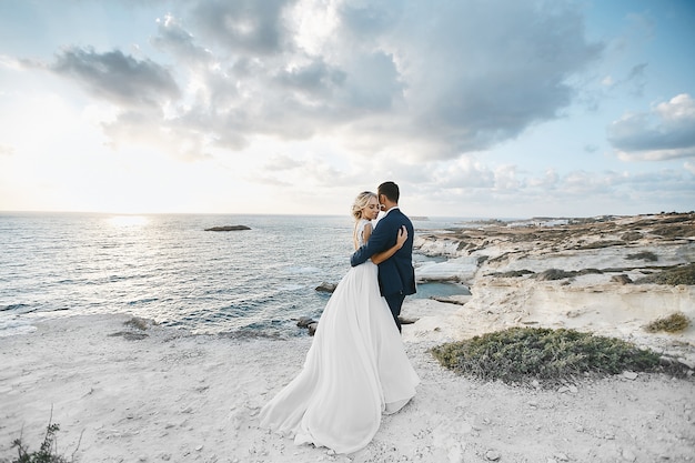 Newlywed couple hugging together on the white rocks at the seacoast at Cyprus. The bride in a wedding dress the groom in a suit posing with beautiful landscape on the background