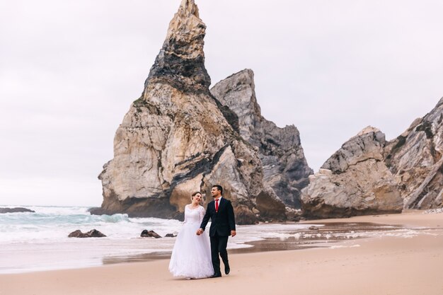 Newlyweds are holding hands and walking on a sandy beach by the ocean and a cliff.