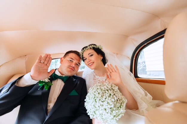 Newlyweds are sitting in the car waving their hands and looking at the camera car interior