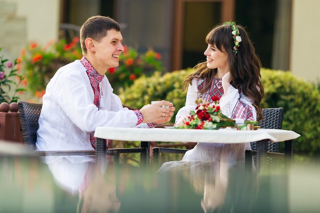 Newlyweds in embroidered clothes in cafe drinking hot coffee and smiling wedding bouquet