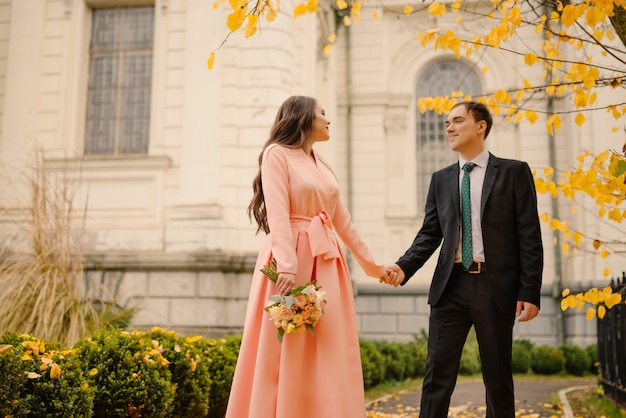 Newlyweds groom and bride walking in autumn park near vintage atmosphere gothic cathedral.