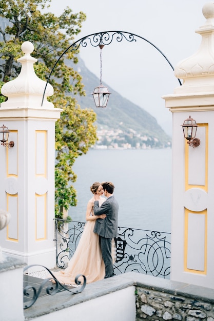 Newlyweds hug and almost kiss under an old arch against the backdrop of lake como