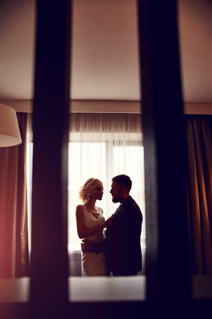 Newlyweds in the morning in the hotel room sitting on the bed hugging and looking at each other in anticipation of the wedding ceremony. Bearded hipster groom and blonde bride in long wedding dress