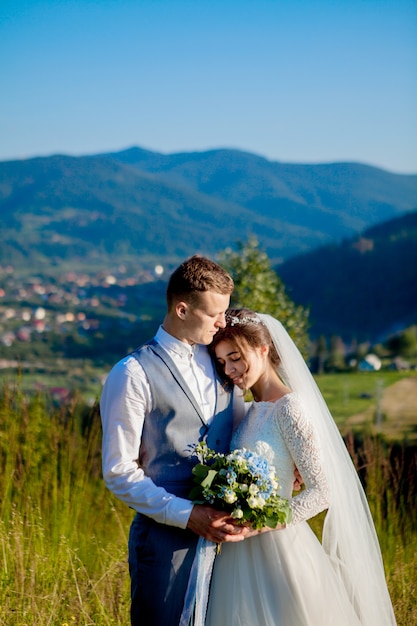 新婚夫婦は山の頂上の草原の中で微笑み、抱き合っています。山の森の中の結婚式の散歩、カップルの優しい感情、バレンタインデーの写真