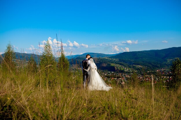 Newlyweds smile and hug each other among the meadow on top of the mountain.