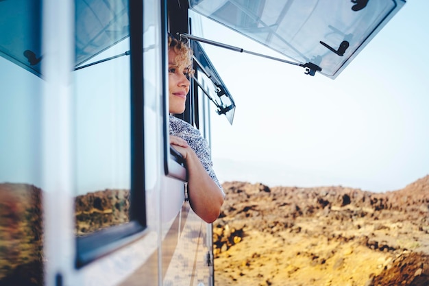 Nice adult woman smile and enjoy freedom at the natural campsite admiring outdoors outside the window of her camper car Concept of summer tourist travel vacation and free female independence people
